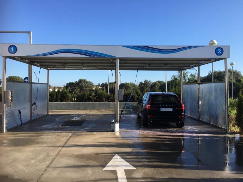 Coche negro en un box de lavado cubierto, con mangueras de agua y paneles laterales dentro de una instalación de autolavado